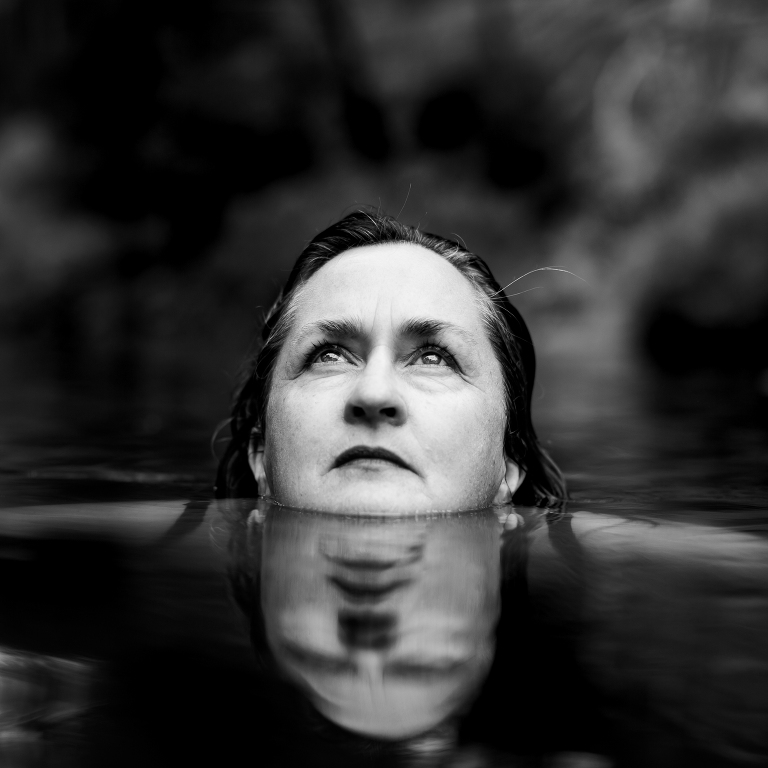 artist Helen Bodycomb in water staring thoughtfully upwards with her face reflected in water