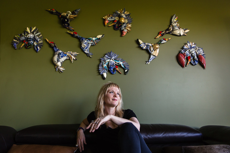 mosaic artist Jessie Yvette Journard Ryan seated on couch smiling into distance with her bird artwork hanging behind her