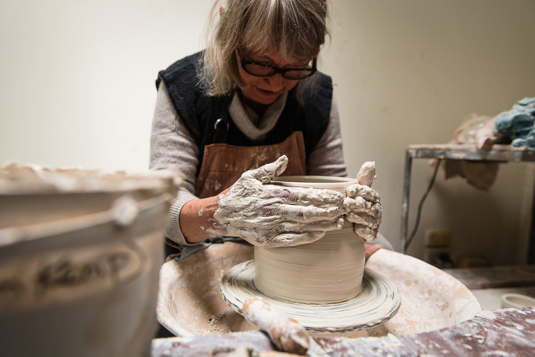 Elderly potter woman Lee Goller turning a pot on her wheel with hands covered in clay
