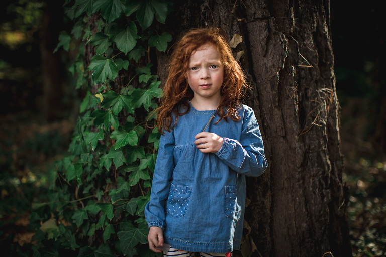 Expressive red haired girl in blue dress facing camera in front of ivy covered pine tree