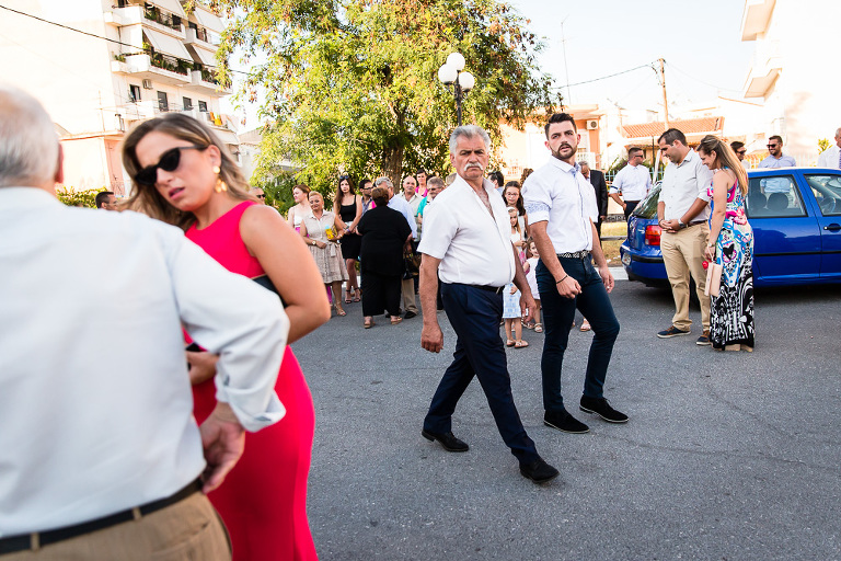 Stern looking older man with adult son walking past wedding guests outside Greek church