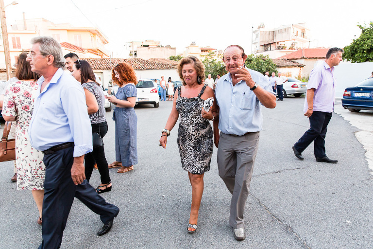 Older man smoking and woman holding his arm walking across car park after Greek wedding