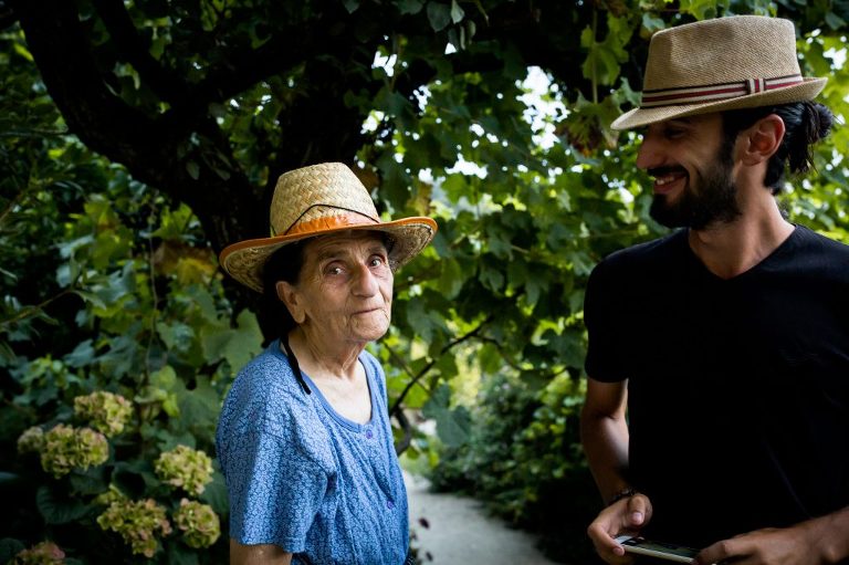 Elderly Albanian woman wearing straw hat while her grandson looks on smiling affectionately