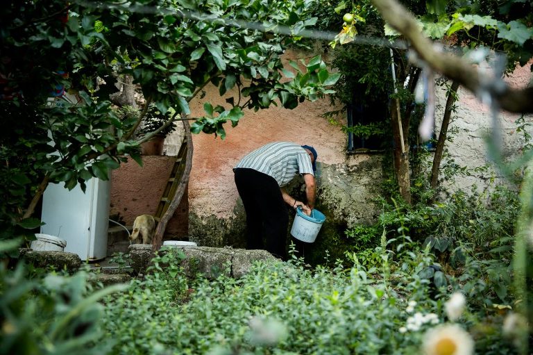 Shepherd bending down cleaning milking bucket at side of old, degraded house surrounded by green garden in Albania