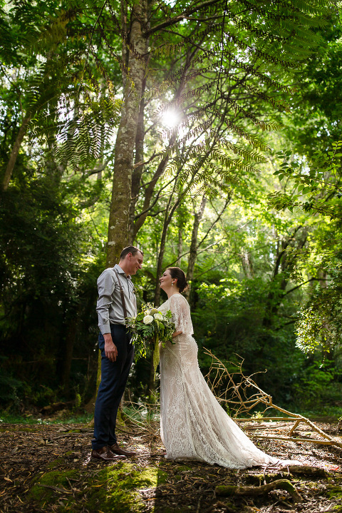bride and groom facing each other in forest with backlight shining through trees